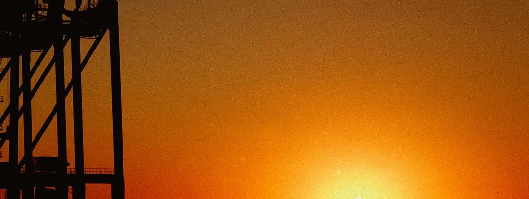 A silhouette of an industrial structure is contrasted against a vibrant sunset, with shades of orange and yellow blending into a warm sky.