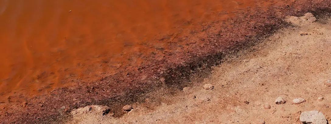 The shoreline of a body of water with deep reddish hues, blending into a lighter sandy bank.