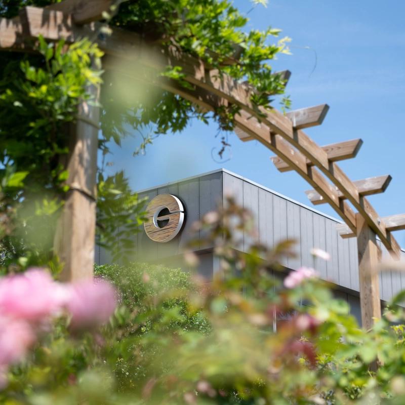 Sunlit garden with blurred pink flowers, a wooden pergola, and the Givaudan Perfumery School beneath a clear blue sky; serene, welcoming mood.