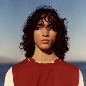 A young person with long, curly hair gazes confidently at the camera, wearing a red sleeveless top over a white shirt.