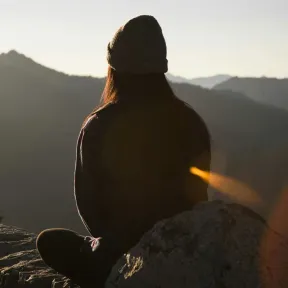 A person sits on a rocky outcrop, looking out over distant mountains at sunset. 