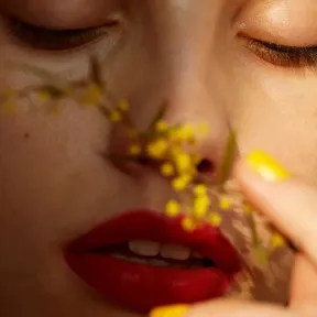 A close-up of a person's face with bold red lips and bright yellow nail polish. Soft sunlight highlights their smooth skin, while a small yellow flower is gently held near their nose, creating a serene and intimate mood.