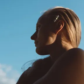 A profile shot of a woman with long, blonde hair adorned with a small white flower, set against a bright blue sky. The image conveys a serene and thoughtful atmosphere.